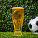 A 16 oz. plastic pilsner glass featuring a FIFA World Cup trophy design, filled with beer and placed on grass with a soccer ball in the background.