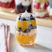 A clear stemless wine tasting glass filled with layered dessert, including yellow custard, blueberries, crumbled cookies, and topped with whipped cream.