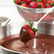 A fresh strawberry being dipped into melted dark chocolate coating with a bowl of strawberries in the background.