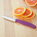 A small paring knife with a white blade and an eggplant purple handle, placed on a wooden cutting board next to sliced oranges.