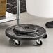 A black round trash can dolly with four swivel caster wheels designed for easy mobility of large containers.
