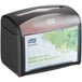 A black tabletop interfold napkin dispenser with a clear front panel displaying a Tork advertisement.