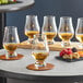 A set of clear Glencairn whiskey glasses, each partially filled with whiskey, displayed on a table with coasters and a small plate of assorted fruits and nuts.