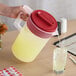 A large plastic pitcher with a red lid being used to pour a yellow beverage into a glass.