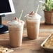 A pair of iced coffee drinks topped with whipped cream and cocoa powder, served in clear plastic cups with striped straws.