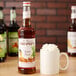 A bottle of Monin Premium Butter Pecan Flavoring Syrup next to a white mug topped with whipped cream.