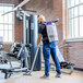 A person using a cordless backpack vacuum to clean the floor of a gym with exercise equipment.