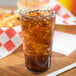 A Carlisle clear plastic tumbler filled with brown liquid and ice on a table with a sandwich and fries.