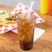 A Carlisle clear plastic tumbler with brown liquid and ice on a table with a straw.