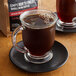 A glass mug of brown liquid on a black plate next to a bag of Crown Beverages Emperor's Finest Whole Bean Decaf Coffee.