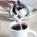 A person pouring milk from a Bon Chef stainless steel server into a cup of coffee.