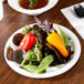 A white paper plate with a salad, small pepper, and bowl of soup on a counter.