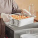 A person in a chef's uniform holding a Vollrath stainless steel steam table pan of food.