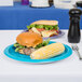 A turquoise blue Creative Converting paper plate with a sandwich and corn on the cob on a table.