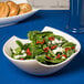 A white porcelain Samurai bowl filled with salad on a counter with bagels and a blue tablecloth.