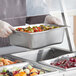 A person in a white uniform holding a Vollrath stainless steel pan of food.