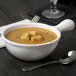 A white Tablecraft cast aluminum soup bowl with a handle filled with soup and a spoon next to it.