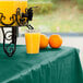 A glass of orange juice sits on a table with a Creative Converting hunter green plastic tablecover next to a drink dispenser and oranges.