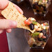 A person holding a chip over a small clear plastic cube bowl filled with food.
