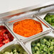 A row of stainless steel Vollrath hexagon pans filled with food including cucumbers, tomatoes, broccoli, and shredded carrots.