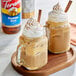 Two mugs of coffee with whipped cream and cinnamon sticks on a table with a bottle of Torani Pumpkin Spice syrup.