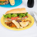 A sandwich and potato chips on a Mimosa yellow Creative Converting paper plate next to a fork and knife.