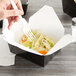 A person's hand holding a fork over a Fold-Pak black paper take-out box of food.