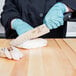 A person in blue gloves using a Mercer Culinary Genesis Santoku knife to cut a piece of meat.