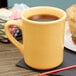 A yellow Tuxton china mug filled with brown liquid on a table with a cup of coffee and a plate of food.