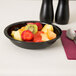 A black Cambro round ribbed bowl filled with fruit on a table with a spoon.