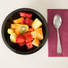 A bowl of fruit in a black Cambro round ribbed bowl on a table with a spoon and a fork.