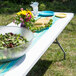 A Flash Furniture white plastic folding table with food and flowers on it.