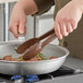 A person using Mercer Culinary brown high temperature plastic tongs to stir food in a pan on a stove.