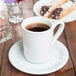 A Tuxton bright white china mug filled with coffee on a saucer on a wooden table with pastries.