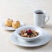 A bowl of cereal and a white Tuxton Alaska mug filled with brown liquid on a table.