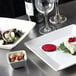 A Tuxton TuxTrendz Santorini square china plate with salad on a table with wine glasses.