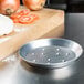 An American Metalcraft heavy weight aluminum pizza pan with perforations on a counter next to tomatoes and a cutting board.