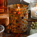 An amber mosaic glass votive candle holder with a lit candle inside.