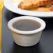 A white Tuxton china ramekin filled with brown liquid on a table with a plate of food.