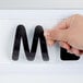 A hand using a black letter to write on a white letter board.