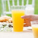 A hand holding a close-up of a WNA Comet Classicware clear plastic fluted tumbler filled with orange juice.