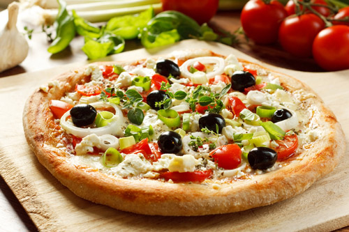 Greek pizza on pizza peel with tomatoes, garlic, and greens in background