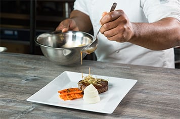 A chef drizzles demi-glace around a plate using a spouted saucier