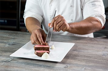 A chef uses precision tongs to plate three pieces of steak next to whipped potatoes