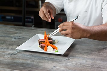 A chef uses precision tongs to plate carrots by artfully leaning them against each other
