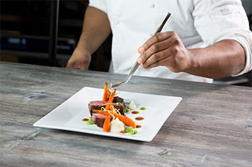 A chef uses precision tongs to garnish a plate with fried leeks