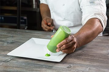 A chef places dots of pea puree around a plate using a large squeeze bottle