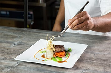 A chef uses precision tongs to garnish a steak with fried leeks