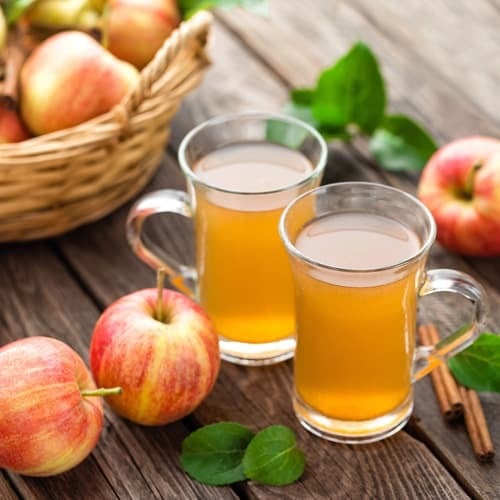 two glasses of apple cider next to two apples and cinnamon sticks on wooden table