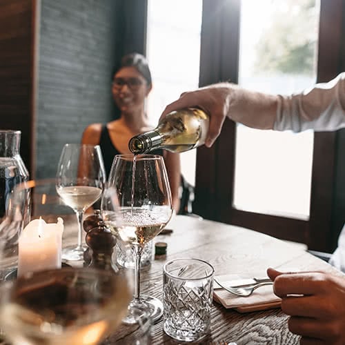 male pours white wine into glass on a candlelit table while female smiles in the background